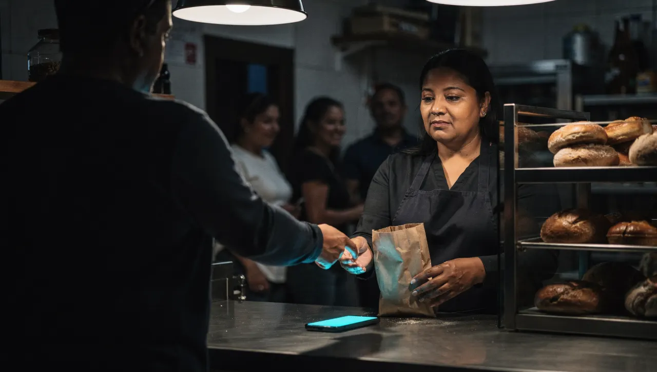La dueña de una panadería atiende a un cliente mientras su celular brilla sobre el mostrador.